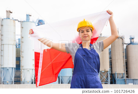 Young female engineer in helmet waving state flag of Poland while standing in front of big tanks at chemical plant 101726426