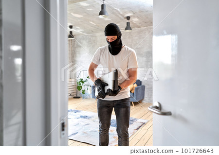 Masked face. Man burglar stealing tv set from house. Thief with black balaclava stealing modern expensive laptop from apartment.  101726643