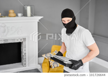 Thief with black balaclava stealing modern expensive laptop from apartment. Masked face. Man burglar stealing tv set from house Thief with black balaclava stealing modern expensive laptop from apartment. Masked face. Man burglar stealing tv set from house 101726644