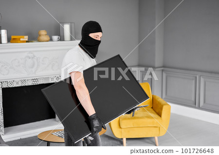 Thief with black balaclava stealing modern expensive television. Masked face. Man burglar stealing tv set from house Thief with black balaclava stealing modern expensive television. Masked face. Man burglar stealing tv set from house 101726648