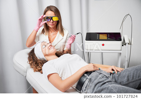 Female beautician in protective glasses using diode laser device while performing skin rejuvenation procedure in beauty salon. Young woman in silver goggles lying on daybed and smiling. 101727824