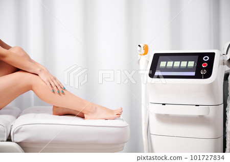 Cropped photo of beautiful female legs after laser hair removal procedure. On white background slender naked legs on couch next to laser hair removal machine. Aesthetic medicine concept. 101727834