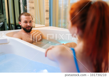 Close-up view from back of unrecognizable redhead young woman wearing bikini talking with handsome bearded man sitting in double hot bathtub with hydromassage, at luxury spa salon, enjoying weekend. 101730210
