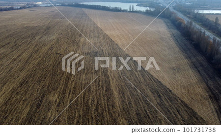 Large plowed field with plowed land and yellow dry straw after harvesting wheat, a lake, a highway with driving cars on an autumn day. Agro industrial agricultural farm landscape. Aerial drone view. Large plowed field with plowed land and yellow dry straw after harvesting wheat, a lake, a highway with driving cars on an autumn day. Agro industrial agricultural farm landscape. Aerial drone view. 101731738
