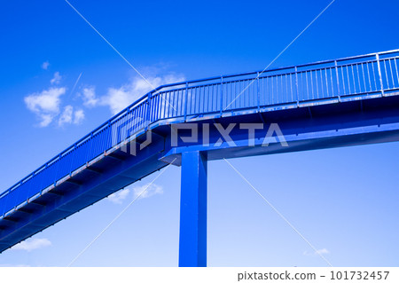 A modern pedestrian overpass across a highway. Blue bridge without people against blue sky in summer day. Minimalistic architecture, urban infrastructure. Bridge on stilts high above a ground. 101732457
