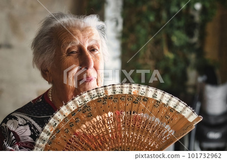 An old woman with white hair indoors waving a traditional wooden fan to keep herself cool in the summer 101732962