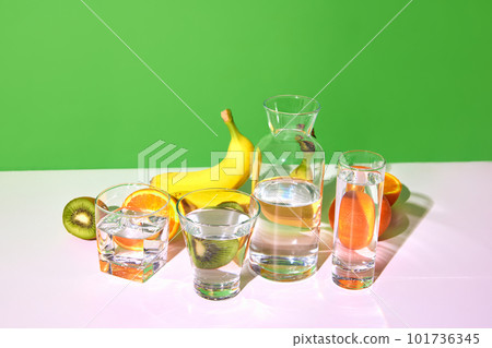 Nutrients, vitamins. Still life with fresh fruits kiwi, banana, orange and glasses with water on table over green studio background. Balanced diet. Bright colors 101736345