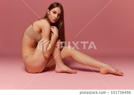 Portrait of young girl posing in beige underwear sitting on floor, posing against pink studio background Portrait of young girl posing in beige underwear sitting on floor, posing against pink studio background 101736498