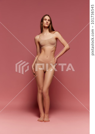 Full-length portrait of young beautiful girl with slim, fit body shape, posing in beige underwear against pink studio background 101736545