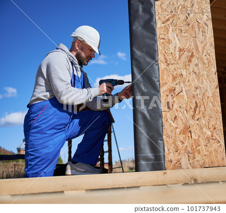Male builder installing black corrugated iron sheet used as facade of future cottage, blue sky on background. Man worker building wooden frame house. Carpentry and construction concept. Male builder installing black corrugated iron sheet used as facade of future cottage, blue sky on background. Man worker building wooden frame house. Carpentry and construction concept. 101737943