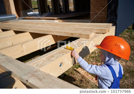 Boy toddler playing as builder on construction site. Child carpenter in orange helmet and blue overalls learning to build wooden frame house outdoor on sunny day. Carpentry and workshop concept. 101737966