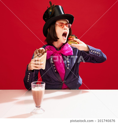 Stylish woman in cylinder hat and tailcoat with pink bow eating burger and chocolate sticks against red background. Sweet and salt Stylish woman in cylinder hat and tailcoat with pink bow eating burger and chocolate sticks against red background. Sweet and salt 101740876