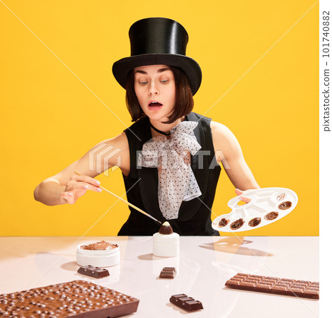 Woman, in image of food painter, in cylinder hat making chocolate sweets, painting candies against yellow background 101740882