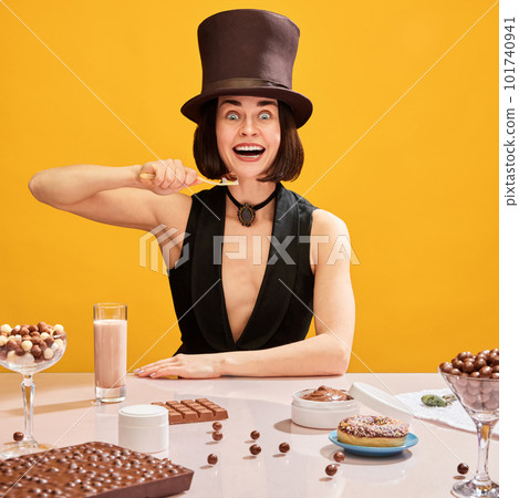 Crazy looking, smiling woman in cylinder brushing teeth with chocolate paste over yellow background. Chocolate sweets all around the table Crazy looking, smiling woman in cylinder brushing teeth with chocolate paste over yellow background. Chocolate sweets all around the table 101740941