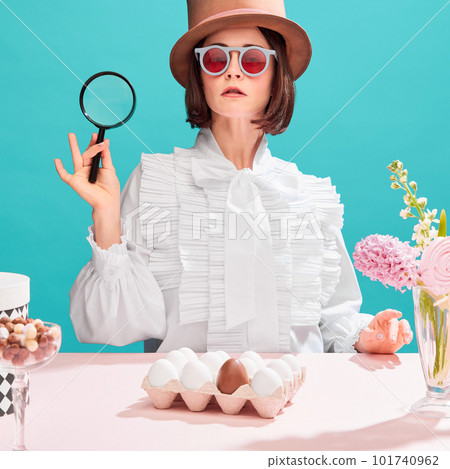 Woman in cylinder hat and sunglasses holding magnifying glass over blue background. Eggs and sweets on table. Holidays preparation 101740962