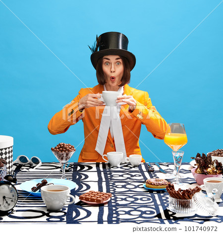 Emotive woman in image of hatter, in top hat and orange jacket drinking coffee over table full of chocolates on blue background 101740972
