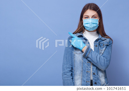 Close up portrait of woman isolated over blue background. Female dresses medical flu mask and gloves, pointing index finger aside. Healthcare, coronavirus, civid19, copy space for advertisment. Close up portrait of woman isolated over blue background. Female dresses medical flu mask and gloves, pointing index finger aside. Healthcare, coronavirus, civid19, copy space for advertisment. 101741651
