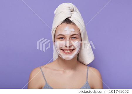 Studio portrait of delighted magnetic young female looking directly at camera, applying mask, caring about skin condition, smiling sicnerely, having wet hair, posing isolated over lilac background. 101741692