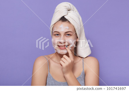 Picture of cheerful tender young girl having spa procedures, wearing pajama, having towel on her wet hair, looking directly at camera, smiling sincerely, applying mask on face. Care concept. 101741697