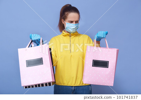 Studio shot of young adorable woman wearing yellow shirt, protective medical mask, disposable gloves from coronavirus, holding bags for shopping in both hands. Covid 19, health care concept. Studio shot of young adorable woman wearing yellow shirt, protective medical mask, disposable gloves from coronavirus, holding bags for shopping in both hands. Covid 19, health care concept. 101741837