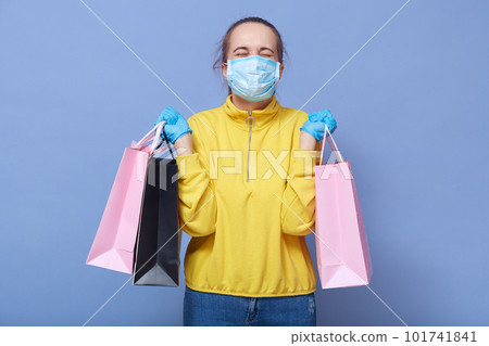 Studio shot of young girl dresses medical mask, protective gloves and casual outfit clutching shopping bags in her hands, keeps eyes closed, looks happy, protects herself while doing shopping. 101741841