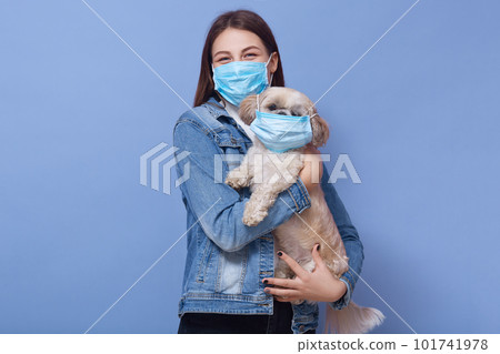 Indoor shot of woman dresses denim jacket protective flumask holds dog pet in medical mask. Coronavirus disease dangerous for people and pets. Coronavirus, pandemic, qaurantine, precautions concept. Indoor shot of woman dresses denim jacket protective flumask holds dog pet in medical mask. Coronavirus disease dangerous for people and pets. Coronavirus, pandemic, qaurantine, precautions concept. 101741978