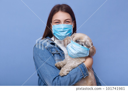 Close up portrait of young woman with long hair, dresses denimjacket and medical mask as precaution against spread of virus, holding her dog in hands with fluemask too. Coronavirus pandemic concept. 101741981