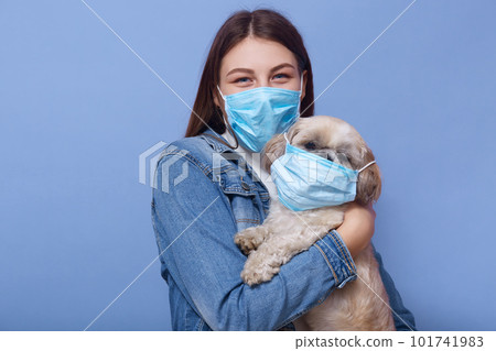 image of dark haired female with dog in hands, lady wearing face mask on her pet for fun, posing isolated over blue studio background, protects from virus. Corona virus, covid19, quarantine concept. image of dark haired female with dog in hands, lady wearing face mask on her pet for fun, posing isolated over blue studio background, protects from virus. Corona virus, covid19, quarantine concept. 101741983