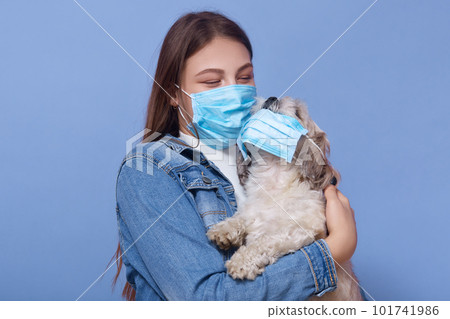 Close up portrait of girl dresses medical mask, holding dog in a protective mask, isolated over blue background. Acute respiratory viral infection. Epidemic precautions, coronavirus, covid 19. Close up portrait of girl dresses medical mask, holding dog in a protective mask, isolated over blue background. Acute respiratory viral infection. Epidemic precautions, coronavirus, covid 19. 101741986