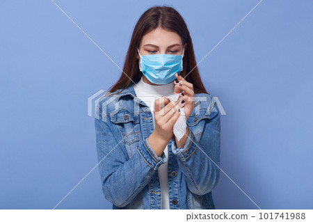 Indoor shot of woman wearing stylish demin jacket and medical blue mask cleaning her hands with wipe isolated over blue background in studio. Coronavirus, covid 19, quarantine, health care concept. 101741988