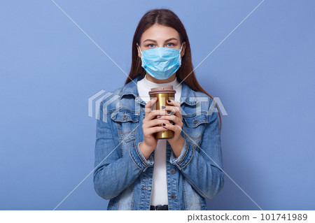 Close up portrait of young Caucasan woman wearing denim jacket, white shirt and blue madical protect mask, holding coffee to take away and looking directly at camera. Coronavirus. covid 19 concept. 101741989