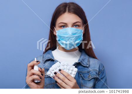 Close up portrait of young brunette wearing antibacterial mask, looking directly at camera, holding pills and nasal spray, wearing jeans jacket, being ill. People and health problems concept. Close up portrait of young brunette wearing antibacterial mask, looking directly at camera, holding pills and nasal spray, wearing jeans jacket, being ill. People and health problems concept. 101741998