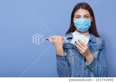 Horizontal shot of young woman dresses medical protective mask, posing in studio background, girl looking with frightened eyes, fear of epidemic, pointing aside with thumb. Coronavirus concept. 101741999
