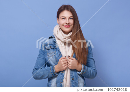 Close up portrait of happy woman wearing denim jacket and scarf, posing isoalted over blue background, lady with positive facial expression, attractive girl looking directly at camera. People concept. 101742018