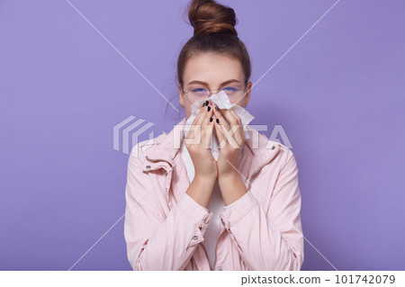 Horizontal shot of young female wearing pale pink jacket and glasses, has hair bun, looking at camera, using napkins for runny nose, posing isolated over lilac studio background. Healthcare concept. Horizontal shot of young female wearing pale pink jacket and glasses, has hair bun, looking at camera, using napkins for runny nose, posing isolated over lilac studio background. Healthcare concept. 101742079