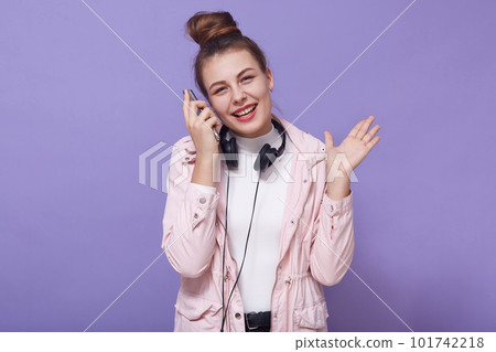 Happy smiling student girl wearing pale pink jacket, white shirt, stands with hair bun, holding smart phone near her ear, listening to favorite music, keeps palm up, looking at camera and laughing. Happy smiling student girl wearing pale pink jacket, white shirt, stands with hair bun, holding smart phone near her ear, listening to favorite music, keeps palm up, looking at camera and laughing. 101742218
