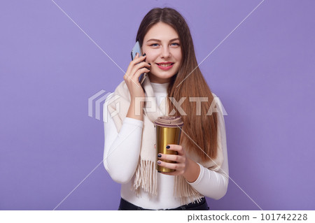 Photo of beautiful girl isolated over lilac background, holding thermo mug in hands, enjoying coffee or tea while talking via smartphone, smiles and looking at camera, wearing casul outfit and scarf. Photo of beautiful girl isolated over lilac background, holding thermo mug in hands, enjoying coffee or tea while talking via smartphone, smiles and looking at camera, wearing casul outfit and scarf. 101742228