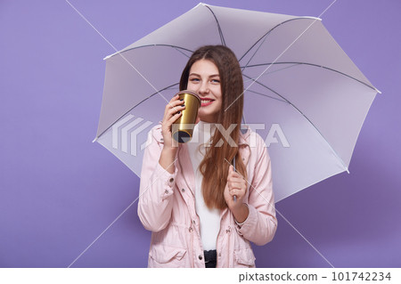 Picture of pleasant good looking cute female holding cup with beverage and umbrella, looking directly at camera, smiling sincerely, drinking hot tea, standing isolated over lilac background. 101742234