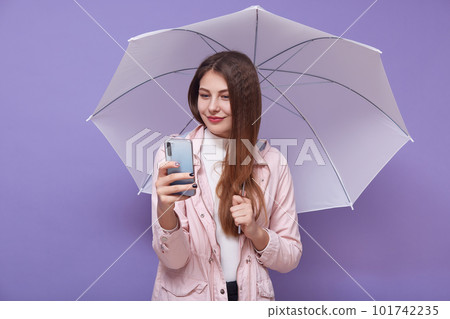 Image of beautiful tender attractive young woman making selfie, talking over videocall, holding smartphone and umbrella, having long hair, bad weather, raining. People and technology concept. 101742235