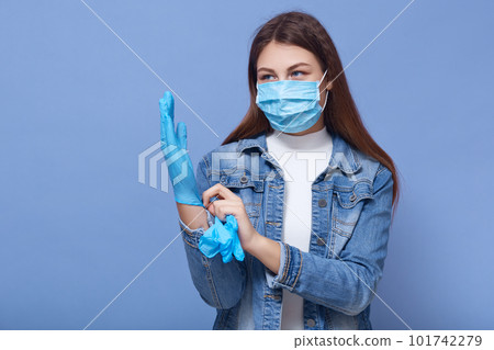 Image of darkhaired Caucasian woman with long hair, wearing medical face mask and putting on disposable gloves, looks at distance, standing isolated over blue background. Coronavirus, covid 19 concept Image of darkhaired Caucasian woman with long hair, wearing medical face mask and putting on disposable gloves, looks at distance, standing isolated over blue background. Coronavirus, covid 19 concept 101742279