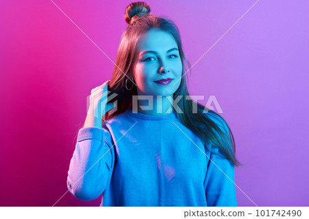 Picture of teenager girl standing and touching her long beautiful hair, has hair bun, dresses casually, posing isolated over pink neon background, looking at camera, looks confident. People concept.. 101742490