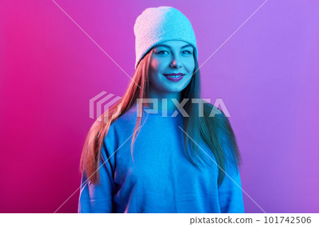 Indoor shot of smiling teenager girl wearing sweater and cap, looking directly at camera with confident facial expression. having long beautiful hair, standing against pink neon wall. People concept. 101742506