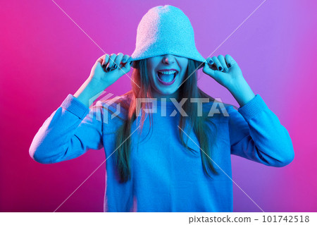 Close up portrait of young woman screaming with cap over her eyes and hands on it, lady posing isolated over pink neon background, female wearing casual sweater. People emotions, lifestyle concept. 101742518