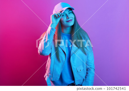 Indoor shot of young beautiful hipster woman in street style fashion, lady wearing hat cap and stylish jacket posing isolated over pink neon light, girl keeping finger on cap's visor. People concept. Indoor shot of young beautiful hipster woman in street style fashion, lady wearing hat cap and stylish jacket posing isolated over pink neon light, girl keeping finger on cap's visor. People concept. 101742541