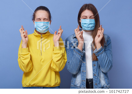 Close up portrait of two young women with crossed fingers, wearing protective flumask, preventing virus infection, females wear casual clothing, posing isolated over blue background. Covid 19 concept. Close up portrait of two young women with crossed fingers, wearing protective flumask, preventing virus infection, females wear casual clothing, posing isolated over blue background. Covid 19 concept. 101742727