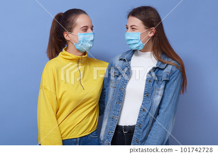 two omen wearing disposable protective face mask and casual outfits. Coronavirus, covid 19, virus epidemic concept, girls friends hugging and looking at each other, isolated over blue background. 101742728