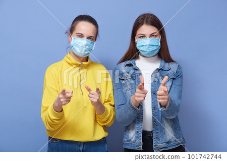 Indoor studio picture of two charismatic good looking females wearing protection masks, making gestures, showing direction with forefingers, standing isolated over blue background. Covid 19 concept. 101742744