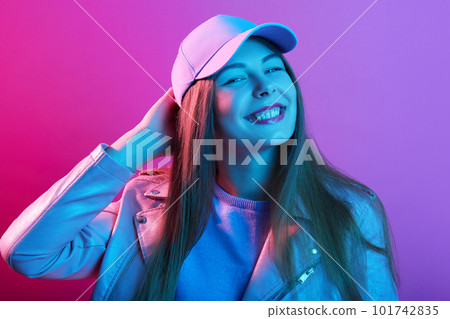 Fashionable woman stands smiling, having fun in stylish outfit. Happy beautiful girl wearing cap and leather jacket. Cheerful young model touching her head and lookins smiling directly at camera. 101742835