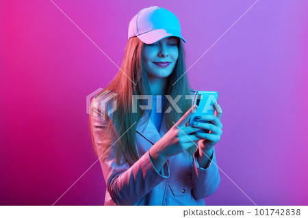 Stylish studio portrait of stylish girl wearing jacket and cap, woman holding phone in hands, checking social networks, posing isolated over pink neon background. Modern technology and people. Stylish studio portrait of stylish girl wearing jacket and cap, woman holding phone in hands, checking social networks, posing isolated over pink neon background. Modern technology and people. 101742838