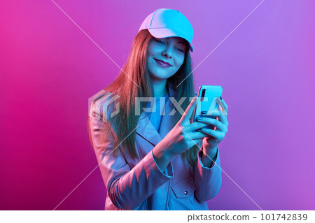 Portrait of young beautiful fashionable model wearing leather baker and baseball cap, holding smart phone in hands, texting with friends or checking social networks. Portrait of young beautiful fashionable model wearing leather baker and baseball cap, holding smart phone in hands, texting with friends or checking social networks. 101742839